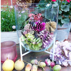 Könnte beinhalten: Ein klares Glasterrarium auf einem weißen Ständer, gefüllt mit grünen und lila Hortensien, Beeren und einem dekorativen Kürbis. Der Text "Hallo HERBST" ist auf dem Glas zu sehen. Gelbe Birnen und Eicheln sind davor angeordnet.