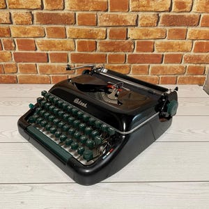 Pu&ograve; includere: Una macchina da scrivere nera vintage con tasti e finiture verdi. La parola "Petrie's" &egrave; visibile sulla macchina da scrivere. La macchina da scrivere &egrave; appoggiata su una superficie di legno chiaro con uno sfondo di muro di mattoni.