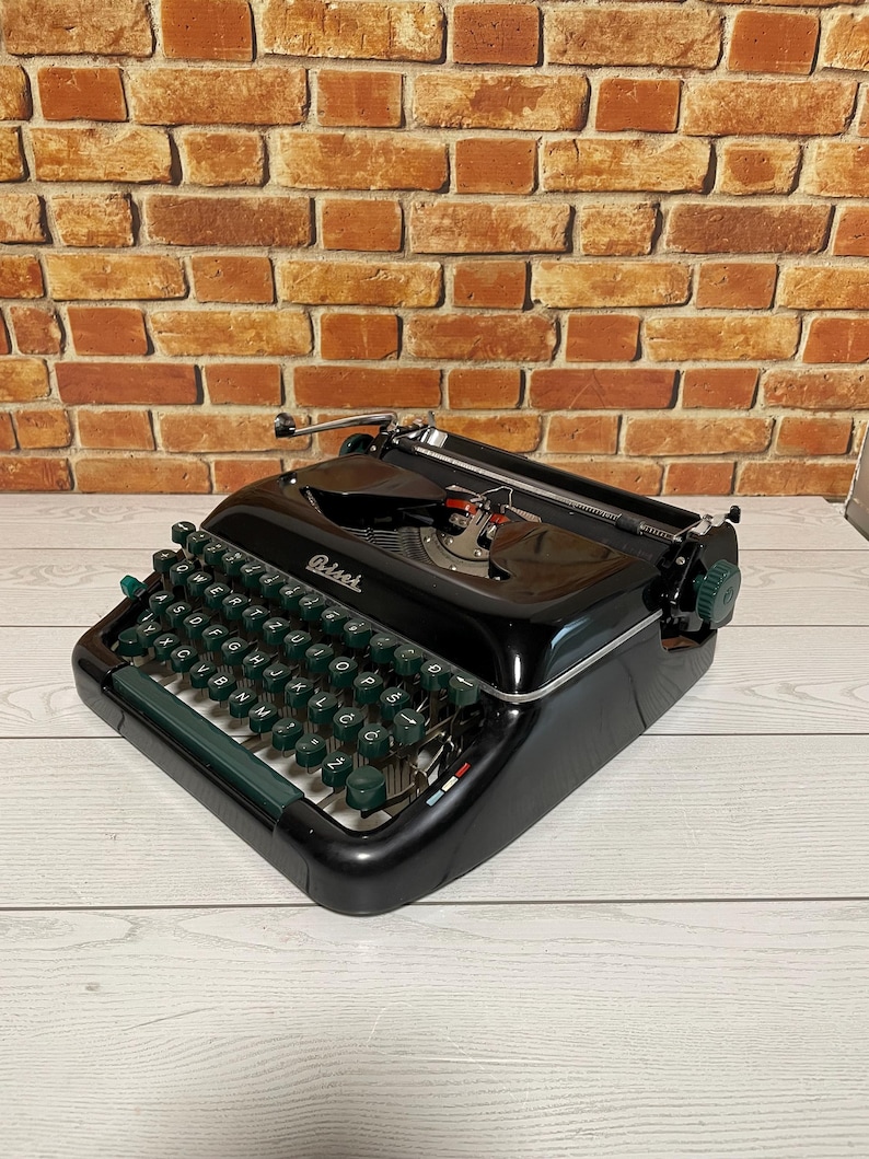 Pu&ograve; includere: Una macchina da scrivere nera vintage con tasti e finiture verdi. La parola "Petrie's" &egrave; visibile sulla macchina da scrivere. La macchina da scrivere &egrave; appoggiata su una superficie di legno chiaro con uno sfondo di muro di mattoni.