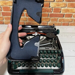 Pu&ograve; includere: Una macchina da scrivere nera vintage con tasti verdi e una copertura nera. Il coperchio &egrave; sollevato, rivelando i meccanismi interni e il nastro rosso. I tasti sono disposti in un layout QWERTY.
