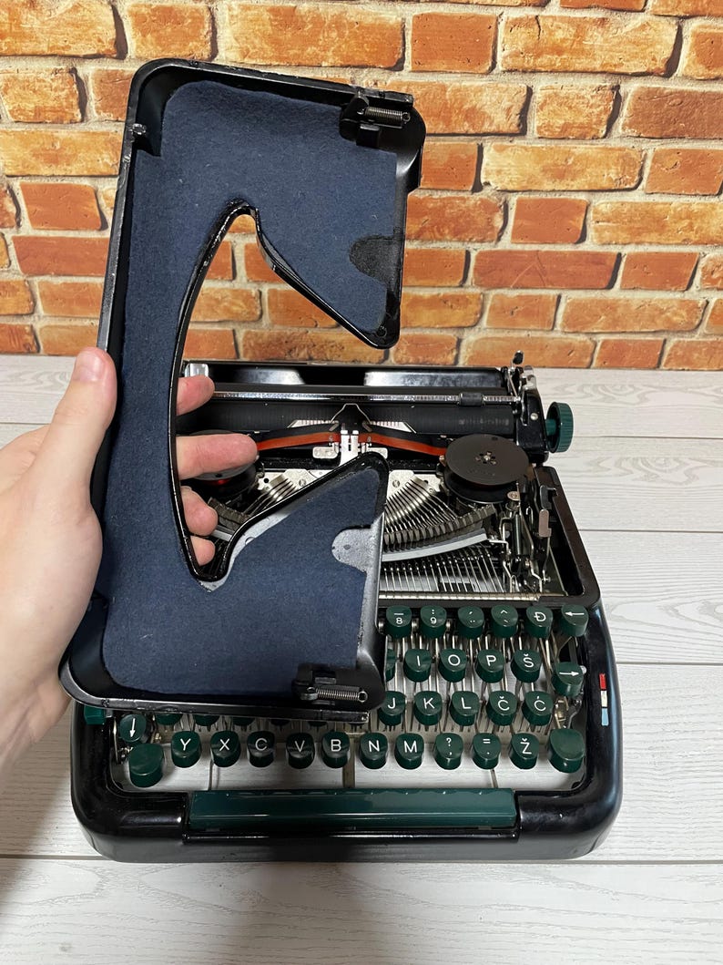 Pu&ograve; includere: Una macchina da scrivere nera vintage con tasti verdi e una copertura nera. Il coperchio &egrave; sollevato, rivelando i meccanismi interni e il nastro rosso. I tasti sono disposti in un layout QWERTY.