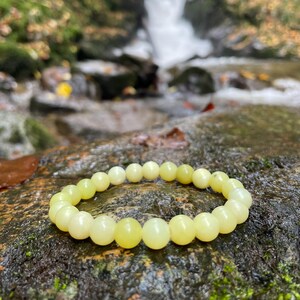 Może przedstawiać: Bransoletka z jasnozielonego jadeitu z okrągłymi koralikami. Bransoletka leży na kamieniu w pobliżu strumienia.
