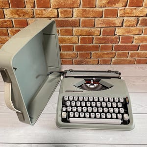 May include: A vintage grey Hermes 3000 typewriter with white keys and a matching carrying case. The typewriter has a classic design with a QWERTY keyboard layout. The case is open, revealing the typewriter ready for use.