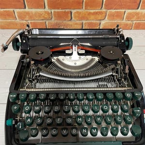 Pu&ograve; includere: Una macchina da scrivere nera vintage con tasti verdi e dettagli cromati. La tastiera ha un layout QWERTY con simboli aggiuntivi. Il nastro &egrave; rosso, su uno sfondo di muro di mattoni.