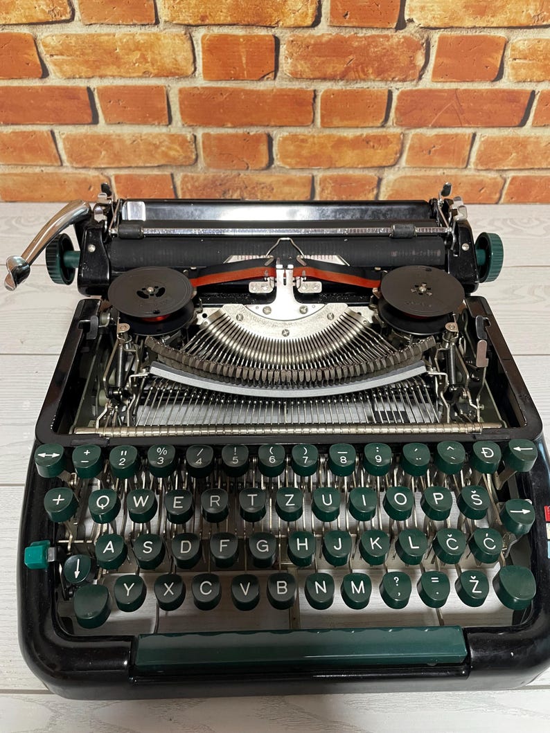Pu&ograve; includere: Una macchina da scrivere nera vintage con tasti verdi e dettagli cromati. La tastiera ha un layout QWERTY con simboli aggiuntivi. Il nastro &egrave; rosso, su uno sfondo di muro di mattoni.