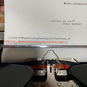 Pu&ograve; includere: Primo piano di una macchina da scrivere vintage con un foglio di carta inserito. La carta mostra testo digitato, inclusi numeri, lettere e simboli. I tasti e i componenti meccanici della macchina da scrivere sono visibili.