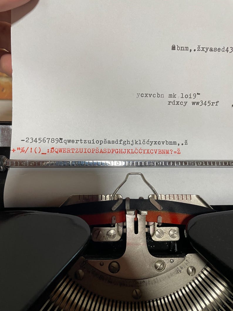 Pu&ograve; includere: Primo piano di una macchina da scrivere vintage con un foglio di carta inserito. La carta mostra testo digitato, inclusi numeri, lettere e simboli. I tasti e i componenti meccanici della macchina da scrivere sono visibili.