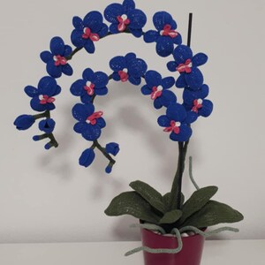 May include: A handcrafted blue crochet orchid plant with pink accents in a burgundy pot. The plant features detailed crochet work, including leaves and stems, and is a decorative item for home decor.