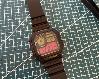 Puede incluir: Reloj digital Casio negro con una esfera rectangular que muestra la hora. El reloj tiene una correa negra y está sobre un fondo de cuadrícula verde. La hora mostrada es 13:07:15.