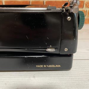 Pu&ograve; includere: Primo piano di una macchina da scrivere vintage nera con dettagli argentati e verdi. Il testo "MADE IN YUGOSLAVIA" &egrave; stampato in oro. La macchina da scrivere &egrave; posta contro un muro di mattoni.