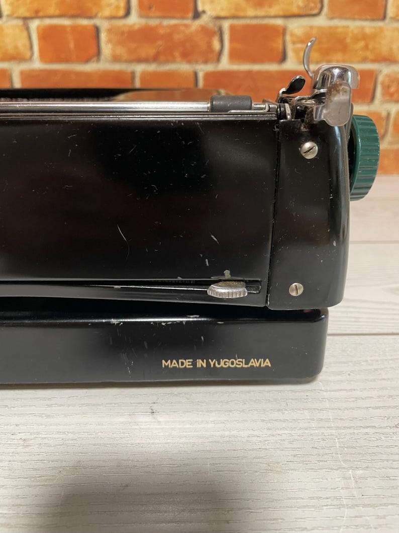 Pu&ograve; includere: Primo piano di una macchina da scrivere vintage nera con dettagli argentati e verdi. Il testo "MADE IN YUGOSLAVIA" &egrave; stampato in oro. La macchina da scrivere &egrave; posta contro un muro di mattoni.