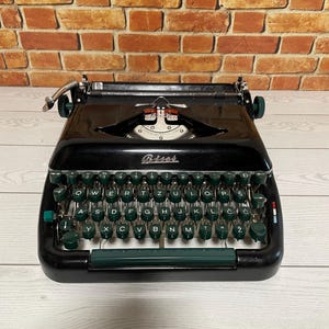 Pu&ograve; includere: Una macchina da scrivere nera vintage con tasti e dettagli verdi. La parola "Biser" &egrave; visibile sulla parte anteriore. La macchina da scrivere &egrave; appoggiata su una superficie di legno chiaro contro un muro di mattoni.