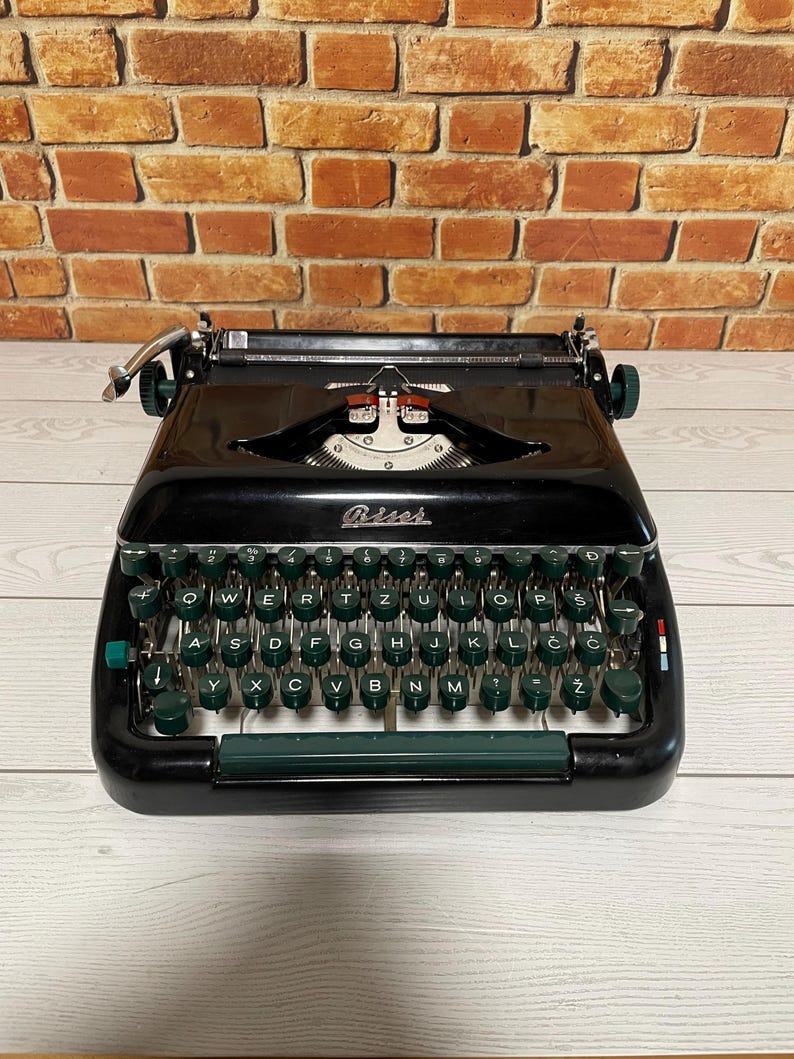 Pu&ograve; includere: Una macchina da scrivere nera vintage con tasti e dettagli verdi. La parola "Biser" &egrave; visibile sulla parte anteriore. La macchina da scrivere &egrave; appoggiata su una superficie di legno chiaro contro un muro di mattoni.