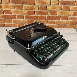 May include: A vintage black typewriter with green keys and accents. The word "Risel" is visible on the front of the typewriter. The keys are arranged in a QWERTY layout. The typewriter sits on a light wood surface.