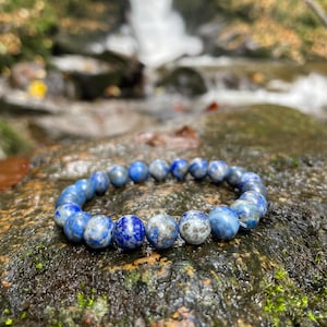 May include: A blue and white gemstone bracelet with round beads. The bracelet is on a mossy rock near a waterfall.