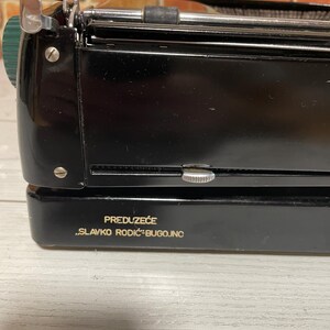 Pu&ograve; includere: Una macchina da scrivere nera vintage con dettagli argentati e verdi. La macchina da scrivere ha una finitura lucida e una prominente leva di ritorno del carrello. Il testo "PREDUZEĆE SLAVKO RODIĆ-BUGOJNO" &egrave; stampato sul davanti.