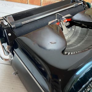 Pu&ograve; includere: Primo piano di una macchina da scrivere nera vintage con tasti argentati e un rullo nero. La macchina da scrivere ha una finitura lucida ed &egrave; ambientata su uno sfondo di muro di mattoni. I tasti sono verdi e la macchina da scrivere &egrave; in buone condizioni.