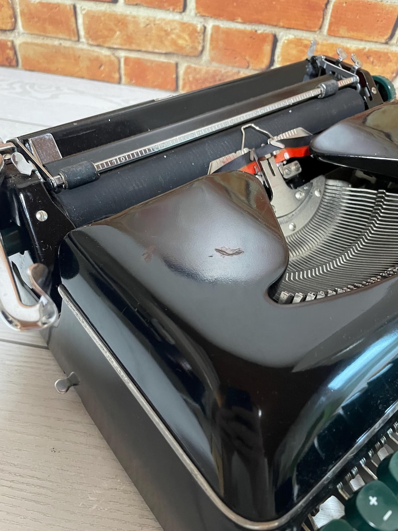 Pu&ograve; includere: Primo piano di una macchina da scrivere nera vintage con tasti argentati e un rullo nero. La macchina da scrivere ha una finitura lucida ed &egrave; ambientata su uno sfondo di muro di mattoni. I tasti sono verdi e la macchina da scrivere &egrave; in buone condizioni.