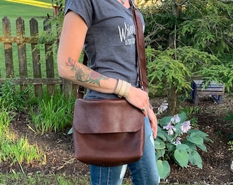 May include: A woman wearing a gray t-shirt with the text "Write as if you mean it" and blue jeans is carrying a brown leather crossbody bag.