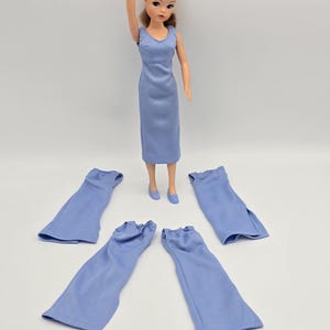 Puede incluir: Una muñeca vintage con un vestido azul claro y zapatos a juego. Cuatro faldas azules claras están dispuestas alrededor de la muñeca. La muñeca tiene el pelo castaño y levanta un brazo.