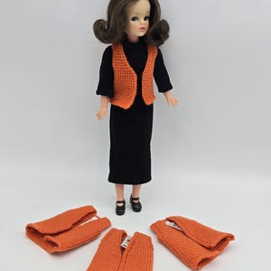 Può includere: Una bambola vintage indossa un vestito nero e un gilet arancione lavorato a maglia. Vengono mostrati tre gilet arancioni aggiuntivi. La bambola ha i capelli castani e indossa scarpe nere. Lo sfondo è bianco.