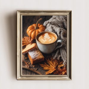 Può includere: Stampa incorniciata con una scena autunnale. Una tazza di caffè con latte art è posta accanto a una piccola zucca, fette di pane e foglie autunnali. Una coperta grigia a maglia aggiunge calore.