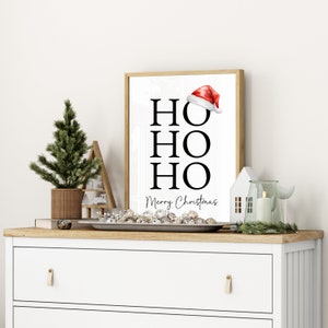 May include: A white framed print with the words "HO HO HO" in black text and "Merry Christmas" in smaller black text. A red Santa hat is above the first "HO". The print is on a white dresser with a small pine tree, a decorative house, and a candle in a glass jar.