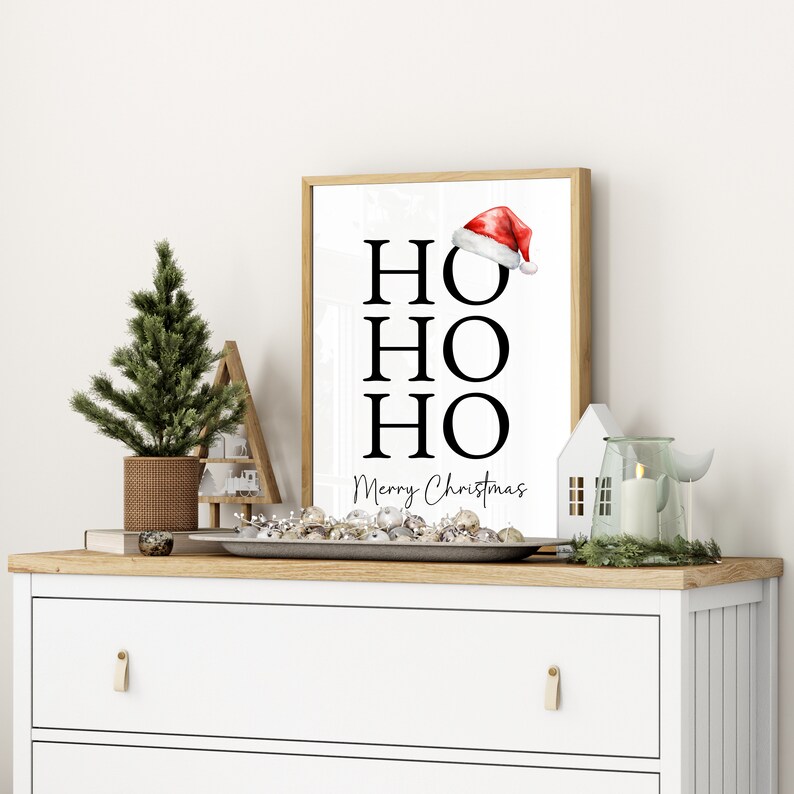 May include: A white framed print with the words "HO HO HO" in black text and "Merry Christmas" in smaller black text. A red Santa hat is above the first "HO". The print is on a white dresser with a small pine tree, a decorative house, and a candle in a glass jar.
