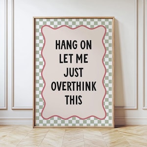 May include: A framed print featuring the phrase "HANG ON LET ME JUST OVERTHINK THIS" in black. The text is on a light pink background with a wavy pink border, set against a green and white checkered pattern. The frame is light brown.