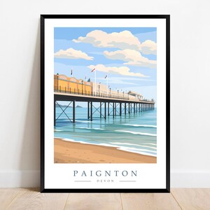 May include: A framed art print of Paignton Pier in Devon, England. The illustration depicts the pier extending over turquoise water, with a sandy beach in the foreground and a blue sky with fluffy clouds above. The text "Paignton Devon" is at the bottom.