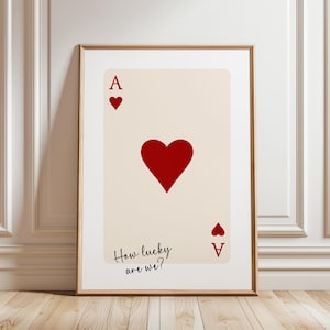 Może przedstawiać: Oprawiony wydruk artystyczny przedstawiający Asa Kier. Karta ma duże czerwone serce na środku, mniejsze serca w rogach i tekst "How lucky are we?" w odręcznej czcionce.