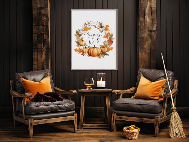 Pode incluir: Uma impress&atilde;o emoldurada com uma coroa de aquarela de folhas de outono e ab&oacute;boras. O texto "Cozy at Home" est&aacute; no centro da coroa. A impress&atilde;o est&aacute; pendurada em uma parede de madeira escura. Duas poltronas de couro com almofadas laranja est&atilde;o em frente &agrave; impress&atilde;o. Uma vassoura est&aacute; encostada na parede &agrave; direita das poltronas.