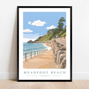May include: A framed art print featuring a coastal scene of Meadfoot Beach in Torquay. The artwork depicts a beach with a sandy shore, a stone wall, and a road along a cliffside. The sky is blue with fluffy white clouds. The print is in a black frame.