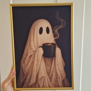 May include: A framed art print featuring a beige ghost figure draped in fabric, holding a black mug with steaming liquid against a dark background. The artwork is enclosed in a thin, metallic gold frame with a rectangular profile and clean edges.