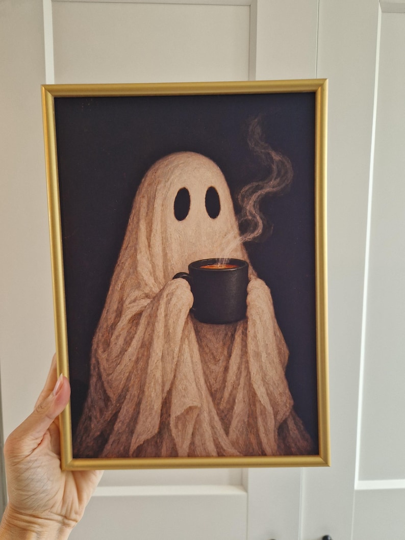 May include: A framed art print featuring a beige ghost figure draped in fabric, holding a black mug with steaming liquid against a dark background. The artwork is enclosed in a thin, metallic gold frame with a rectangular profile and clean edges.