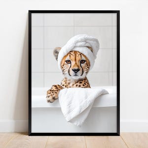 Op de afbeelding: Een ingelijste print met een cheeta jong in een witte badkuip, met een witte handdoek op zijn hoofd. De cheeta heeft bruine vlekken en kijkt de kijker recht aan. Een witte handdoek is over de rand van de badkuip gedrapeerd.