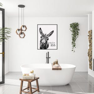 K&ouml;nnte beinhalten: Ein schwarz-wei&szlig; gerahmter Druck eines Esels mit dem Text "nice ass" in einem modernen Badezimmer. Das Badezimmer verf&uuml;gt &uuml;ber eine wei&szlig;e Badewanne, einen Holzschemel und eine H&auml;ngepflanze. Der Raum ist hell und luftig.