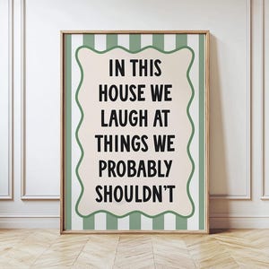 Può includere: Una stampa artistica incorniciata con il testo "IN THIS HOUSE WE LAUGH AT THINGS WE PROBABLY SHOULDN'T". La stampa ha uno sfondo beige con un bordo ondulato ed è ambientata su uno sfondo a righe verdi. La cornice è di colore marrone chiaro.
