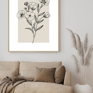 Pode incluir: Um desenho a preto e branco de um ramo de flores sobre um fundo bege, emoldurado. O desenho est&aacute; pendurado numa parede branca acima de um sof&aacute; bege com uma manta e almofadas castanhas.