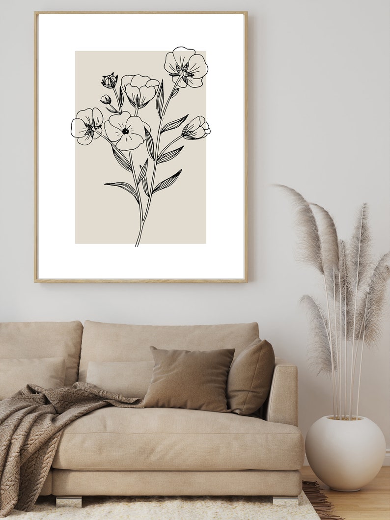 Pode incluir: Um desenho a preto e branco de um ramo de flores sobre um fundo bege, emoldurado. O desenho est&aacute; pendurado numa parede branca acima de um sof&aacute; bege com uma manta e almofadas castanhas.