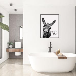 K&ouml;nnte beinhalten: Schwarz-wei&szlig; gerahmter Kunstdruck mit einem Esel und dem Text "nice ASS". Das Kunstwerk wird in einem modernen Badezimmer mit einer wei&szlig;en Badewanne, Kerzen und einer gro&szlig;en gr&uuml;nen Pflanze ausgestellt.