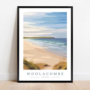 May include: A framed art print of Woolacombe beach in Devon, England. The artwork features a sandy beach, ocean waves, and a blue sky with clouds. The print has a minimalist, vintage travel poster style.