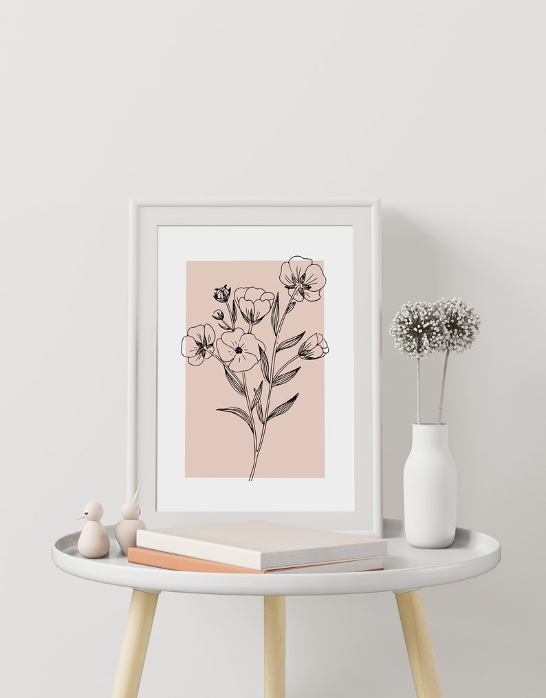 Pode incluir: Uma impress&atilde;o emoldurada branca com um desenho de linha preta de flores sobre um fundo rosa claro. A impress&atilde;o est&aacute; sobre uma mesa auxiliar branca com um livro e duas pequenas figuras de p&aacute;ssaros.