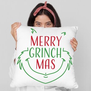 May include: A white pillow with green and red text that reads "Merry Grinch Mas". The text is in a festive, handwritten font. A person is holding the pillow in front of their face, only their eyes are visible.