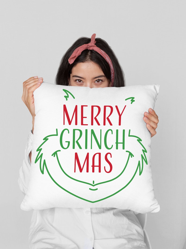 May include: A white pillow with green and red text that reads "Merry Grinch Mas". The text is in a festive, handwritten font. A person is holding the pillow in front of their face, only their eyes are visible.