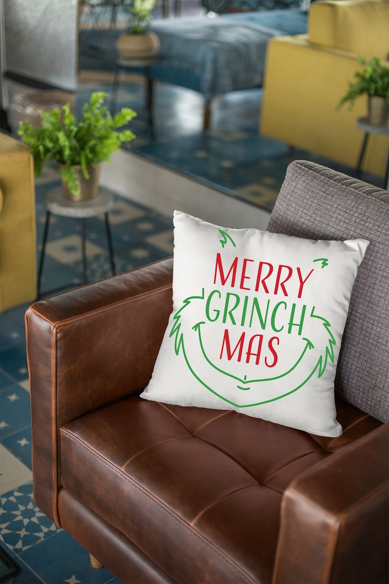 May include: White throw pillow with green and red text that reads "Merry Grinch Mas". The pillow is on a brown leather couch.