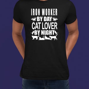 May include: A black t-shirt with white text that reads "Iron Worker By Day Cat Lover By Night". The shirt features a silhouette of an anvil and several cats.