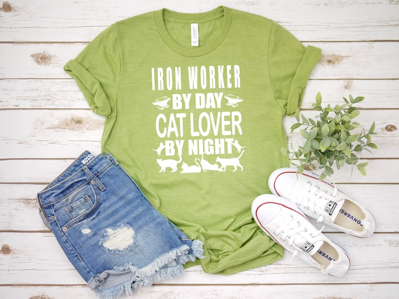 May include: Green t-shirt with white text that reads "Iron Worker By Day Cat Lover By Night". The shirt features a graphic of a wheelbarrow and several cats.