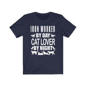 May include: Navy blue t-shirt with white text that reads "Iron Worker By Day Cat Lover By Night". The shirt features a silhouette of a cat and an anvil.