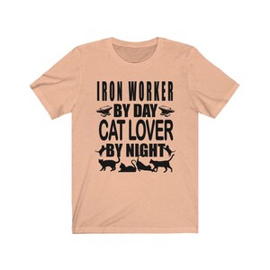 May include: Peach colored t-shirt with black text that reads "Iron Worker By Day Cat Lover By Night". The shirt has a black silhouette of an anvil and four cats.